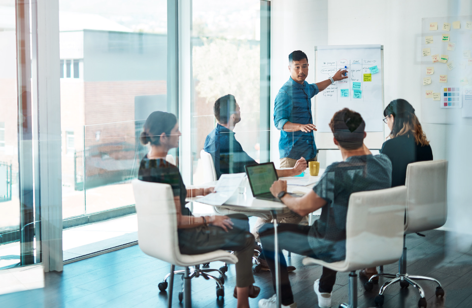 A room of people looking at a speaker who is pointing at a white board of various marketing strategies and tactics to consider. This image represents the header "Key Considerations in Crafting Marketing Strategies & Tactics."